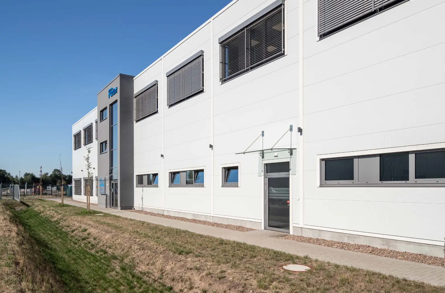 Dargestellt ist ein großes Gebäude, das als Lager- und Fertigungshalle sowie Bürotrakt dienen soll. Hierbei sind ein Personaleingang und ein Haupteingang zu sehen, sowie diverse Fenster im ersten Stock und im Erdgeschoss.
