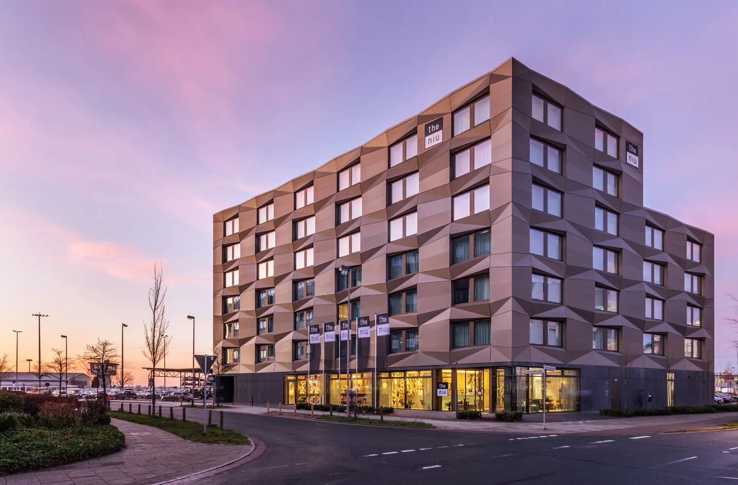 Ein Foto des Hotels "the niu" bei Sonnenuntergang. Es zeigt ein großes Hotel mit eckigen Mustern in der Fassade.  Die untere Etage ist mit einer Glasfront verkleidet und beleuchtet.