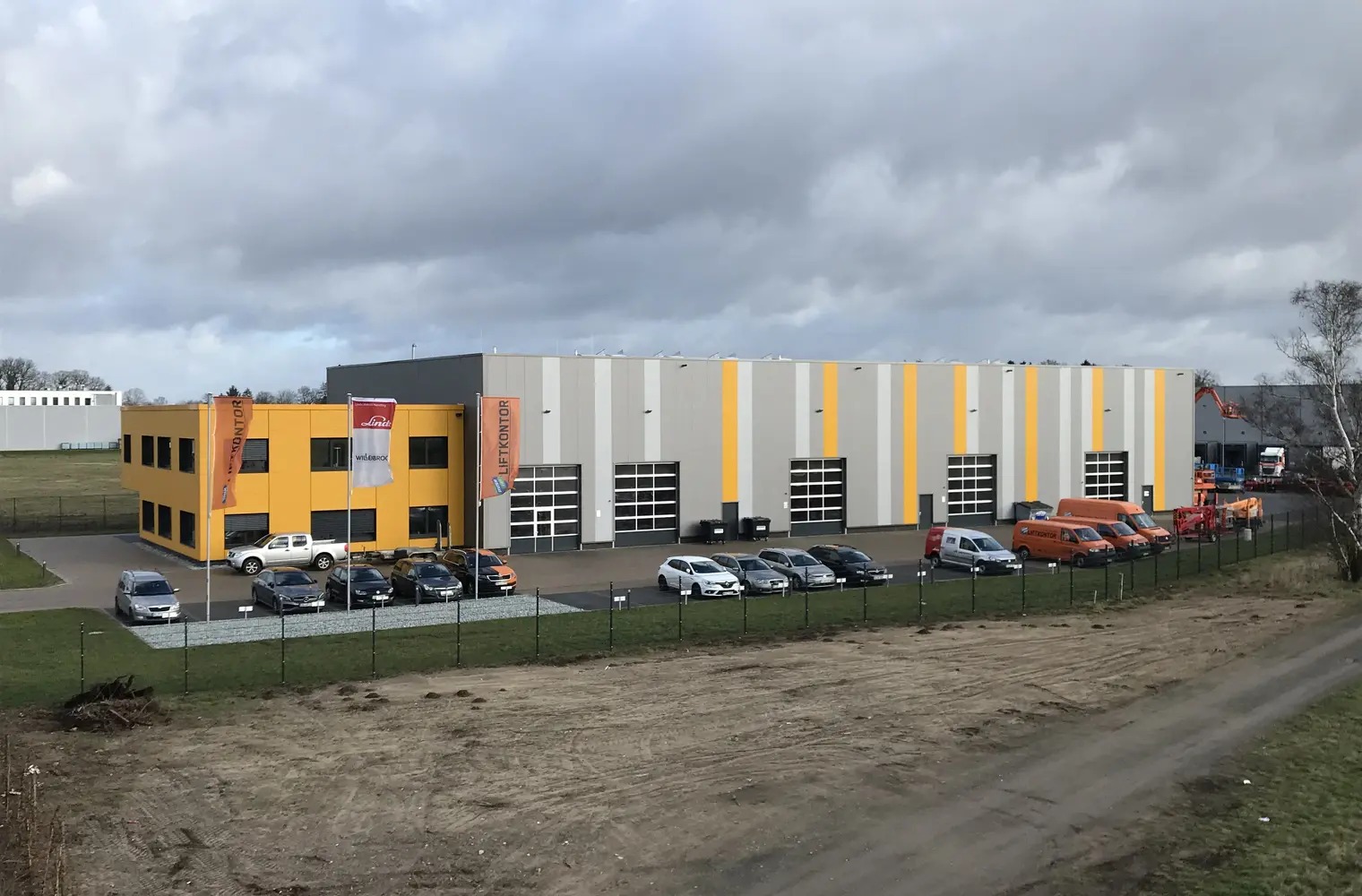Foto der Lager- und Montagehalle "Liftkontor Hannover". Abgebildet ist eine große Halle mit mehreren LKW-großen, halbverglasten Garagen. Davor befindet sich ein Parkplatz, auf dem Pkw abgestellt sind.