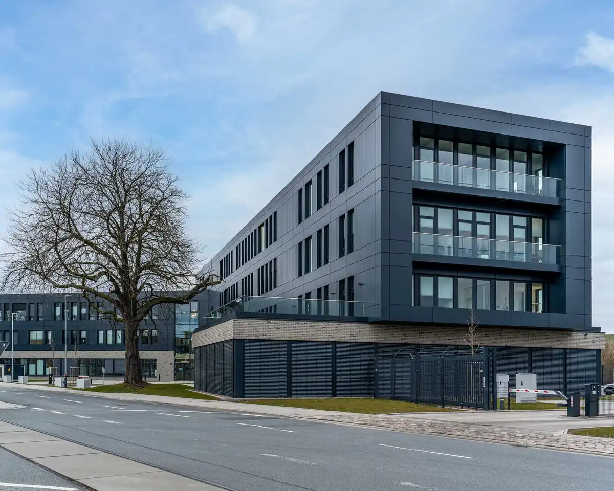 Foto des Bürogebäudes "ADAS". Dargestellt ist ein großes, dunkles Gebäude mit schmalen, bodentiefen Fenstern und einer durchgehenden Fensterfront auf einer Seite. Vor dem Gebäude befindet sich eine Zufahrt zu Parkplätzen.