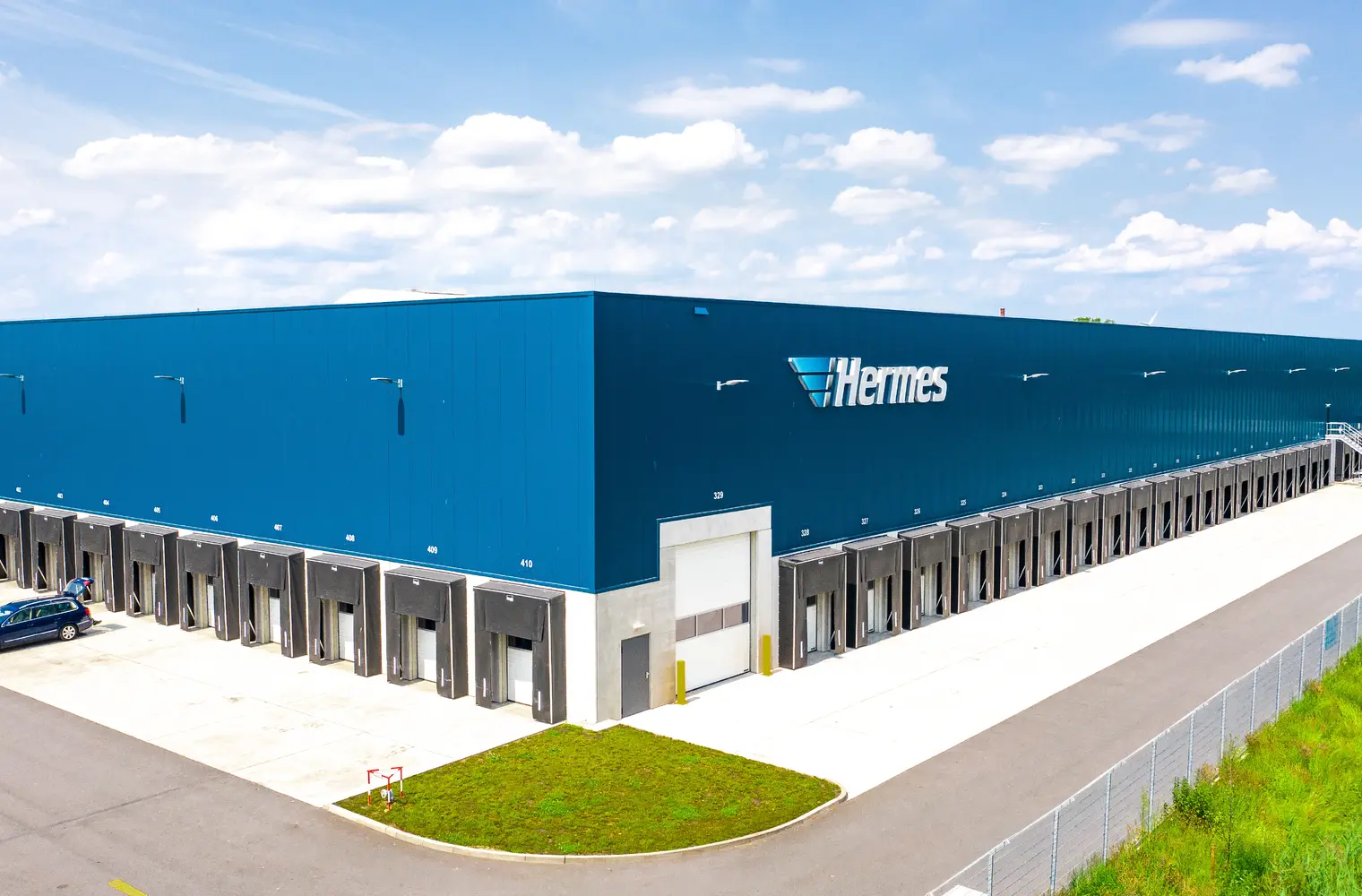Foto des Hochleistungslagers Weyhe - Hermes. Abgebildet ist eine große Warenhalle mit reihenweise Verlademöglichkeiten für Transporter und LKW. Oben das Logo der Firma Hermes mit dem Schriftzug "Hermes".